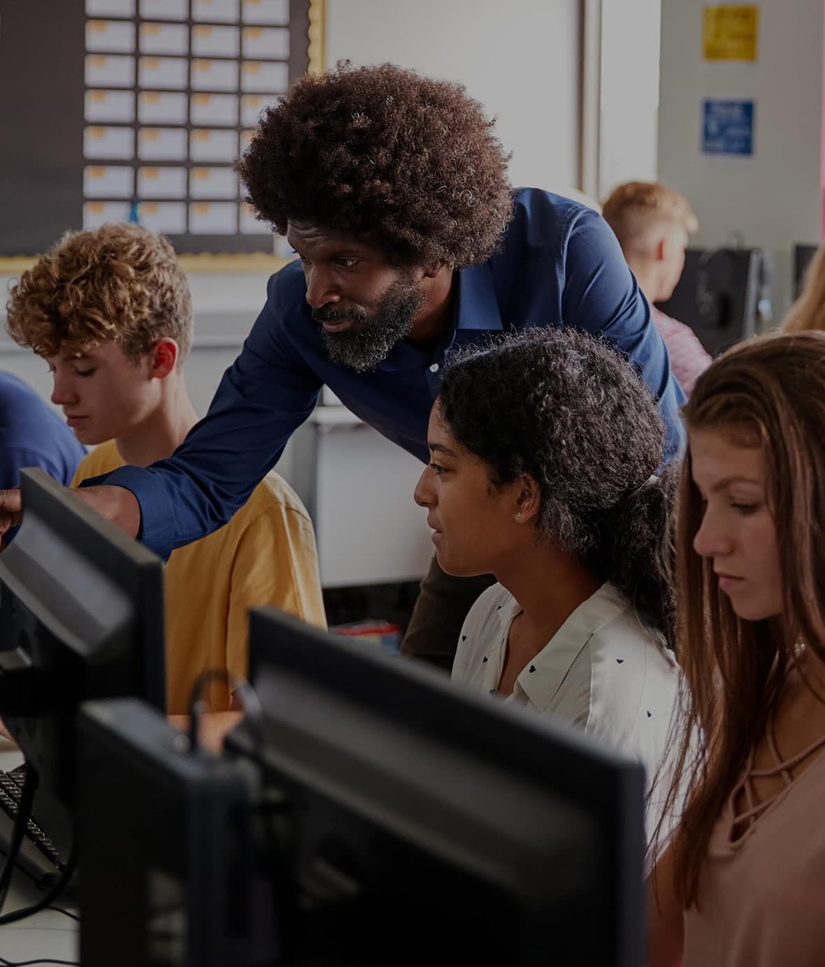 A teacher points at a computer, with students in the background. See how Mojo Helpdesk's education help desk software for schools and universities.
